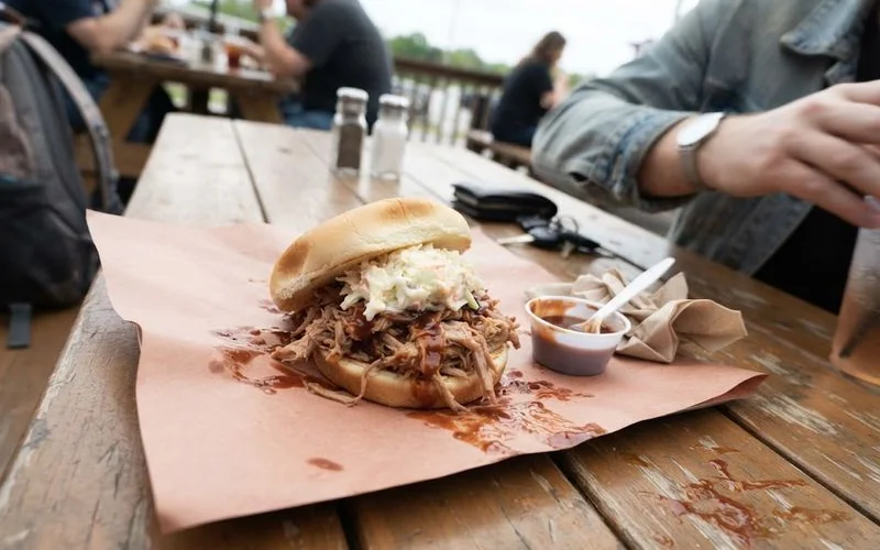 A pulled pork sandwich piled with hand-shredded smoked pork, visible bark mixed in, and a small ramekin of thin vinegar-based sauce on the side